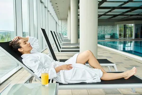 A young, beautiful brunette woman is lounging on a lounge chair next to an indoor swimming pool, basking in relaxation. — Stock Photo