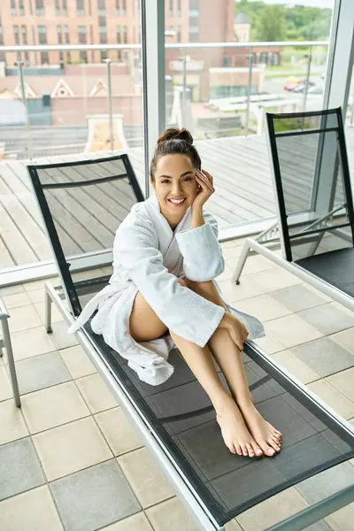 A young, beautiful brunette woman in a bathrobe sits peacefully on a lounge chair in an indoor spa with a swimming pool. — Stock Photo