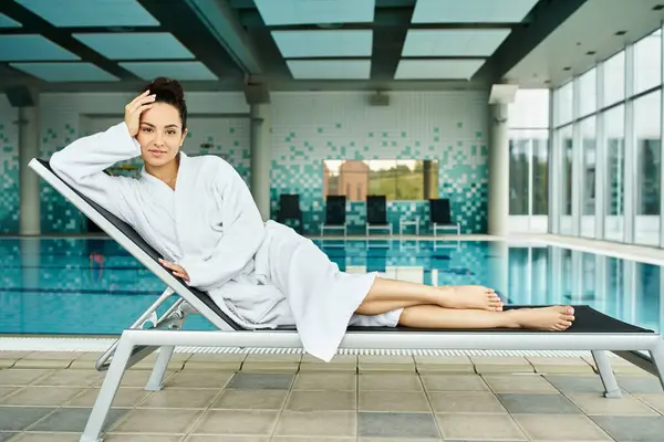 Une jeune femme brune se détend sur une chaise longue à côté d'une piscine intérieure dans un spa luxueux. — Photo de stock