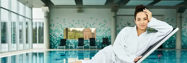 A young brunette woman in a bathrobe sitting serenely by an indoor swimming pool, enjoying a moment of relaxation. — Stock Photo