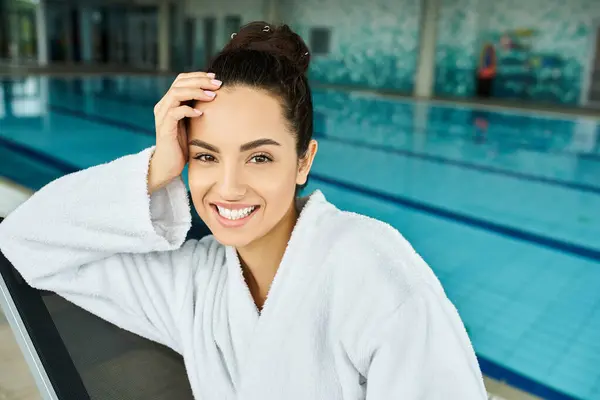 A young and beautiful brunette woman in an indoor spa, sitting gracefully by the inviting swimming pool. — Stock Photo