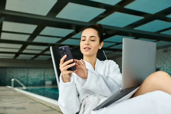 A young, beautiful brunette woman relaxes in a bathrobe by an indoor spa pool, focused on her cell phone. — Stock Photo