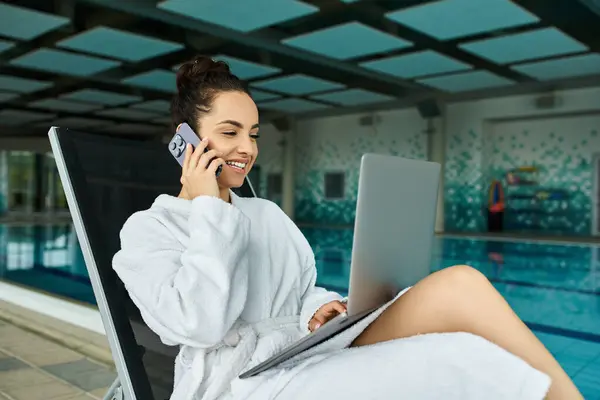 A young, beautiful brunette woman in a bathrobe talking on a cell phone in an indoor spa with a swimming pool. — Stock Photo