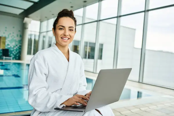 A young, beautiful brunette woman in a bathrobe relaxes by a pool, engrossed in her laptop. — Stock Photo