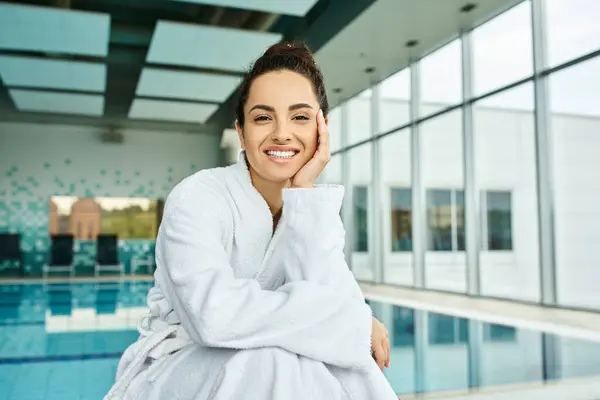 A young brunette woman in a bathrobe enjoying a relaxing moment by the swimming pool at an indoor spa. — Stock Photo