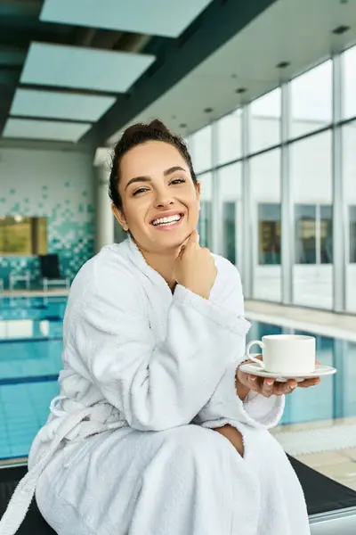 Eine junge brünette Frau genießt einen friedlichen Moment und hält eine Tasse Kaffee in ihrem Bademantel am Innenpool. — Stockfoto