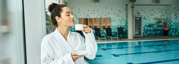 A young brunette woman enjoys a peaceful morning, sipping coffee in a luxurious bathrobe by an indoor spa pool. — Stock Photo