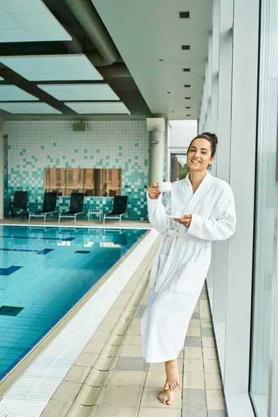 A young, beautiful woman in a bathrobe savoring a cup of coffee, stands beside an indoor swimming pool in a serene spa setting. — Stock Photo