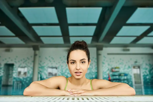 Jeune femme brune en maillot de bain croise les bras alors qu'elle est assise dans la piscine thermale intérieure, respirant tranquillité et paix. — Photo de stock