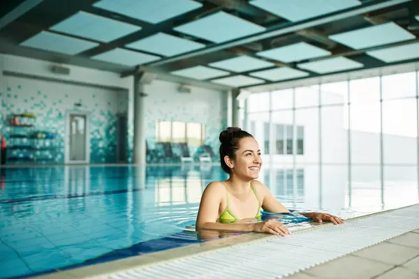 Une jeune, belle femme brune se détend dans un bikini vert dans une piscine spa intérieure, respirant élégance et tranquillité. — Photo de stock