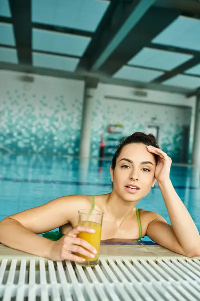 A young brunette woman in a swimsuit enjoys a glass of orange juice while relaxing in an indoor spa swimming pool. — Stock Photo