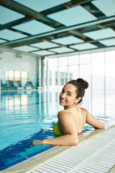 Uma jovem, bela mulher morena em um biquíni verde relaxante na beira de uma luxuosa piscina interior spa. — Fotografia de Stock