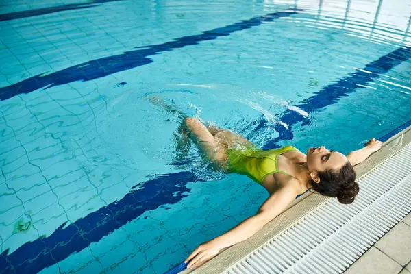 Молода брюнетка в зеленому купальнику насолоджується розслаблюючим плаванням у критому спа-басейні. — стокове фото