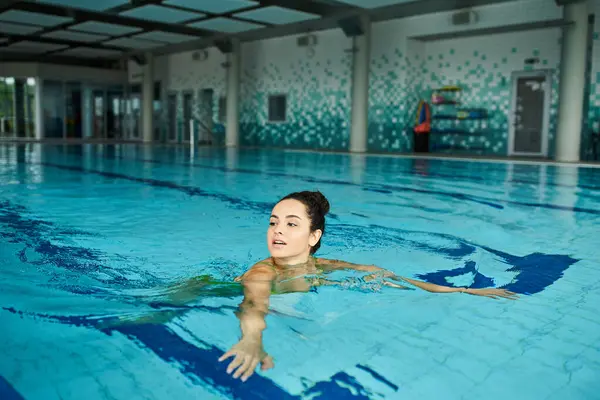 Una joven y hermosa morena nada graciosamente en una tranquila y espaciosa piscina cubierta, encarnando relajación y serenidad.. - foto de stock