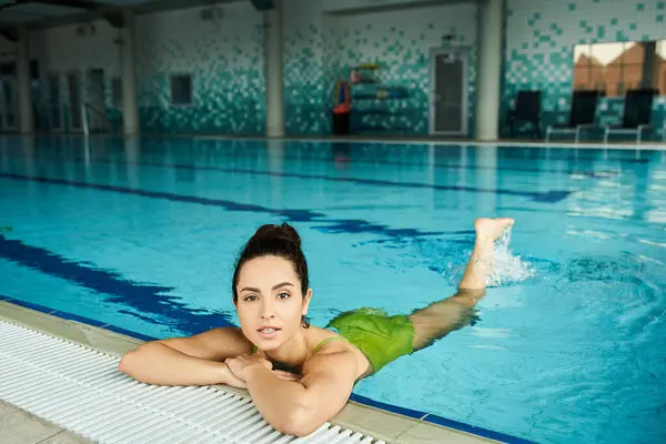 Una joven y hermosa morena en traje de baño verde se relaja en una piscina dentro de un spa interior. - foto de stock