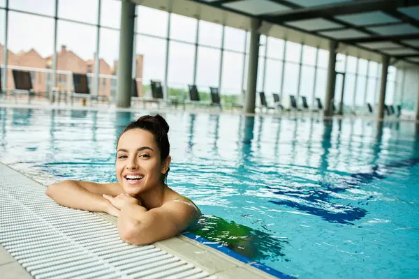 A young brunette woman in a swimsuit smiles as she relaxes in an indoor spa swimming pool. — Stock Photo