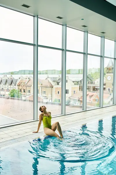 Uma jovem morena de fato de banho verde senta-se graciosamente na beira de uma piscina interior. — Fotografia de Stock