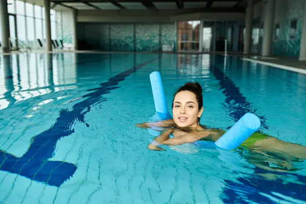 A young, beautiful brunette woman in a swimsuit gracefully swims in a pool with blue rafts under an indoor spa setting. — Stock Photo