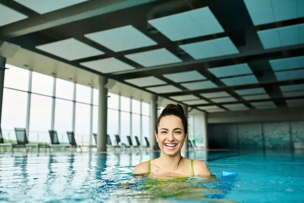A young, beautiful brunette woman in a swimsuit smiles happily while relaxing in an indoor spa swimming pool. — Stock Photo