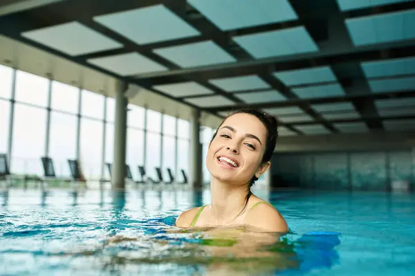 Una joven morena en traje de baño sonríe mientras disfruta de un baño en una piscina cubierta. - foto de stock