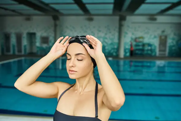 A young woman in a swimsuit and swim cap holding her head in front of an indoor swimming pool. — Stock Photo