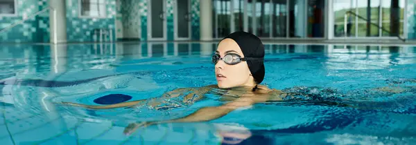 Une jeune femme en maillot de bain et bonnet de bain nageant dans une piscine avec des lunettes, créant des ondulations dans l'eau. — Photo de stock