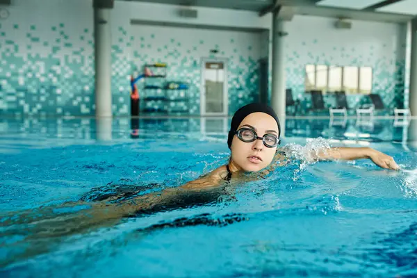 Una mujer joven en traje de baño y gafas nade con gracia en una piscina cubierta, mostrando sus habilidades de natación. - foto de stock