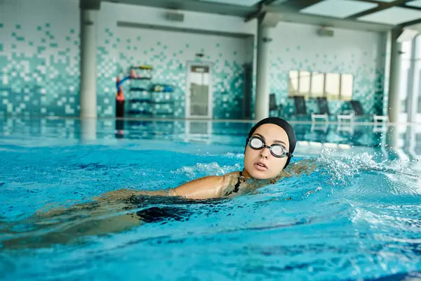 A young woman wearing goggles, a swimsuit, and a swim cap swimming gracefully in an indoor pool. — Stock Photo