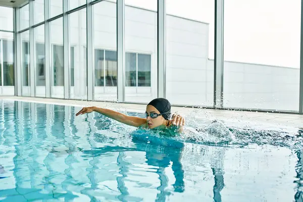 A young woman in a swimsuit and swim cap gracefully swimming in an indoor pool inside a building. — Stock Photo