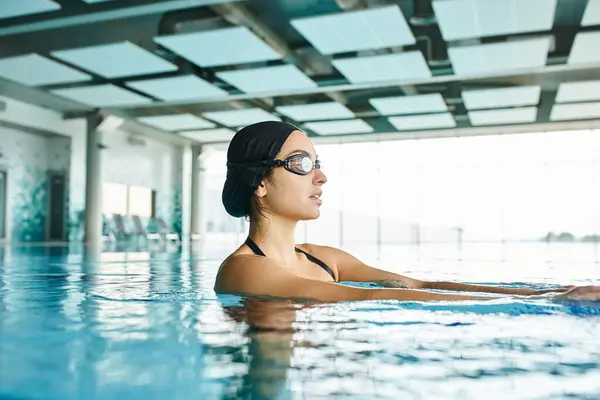 A young woman wearing goggles relaxes in a swimming pool, enjoying a peaceful swim in an indoor spa. — Stock Photo