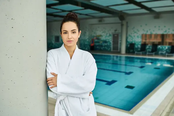 Une jeune femme brune en peignoir se tient gracieusement à côté d'une piscine intérieure, respirant détente et élégance. — Photo de stock