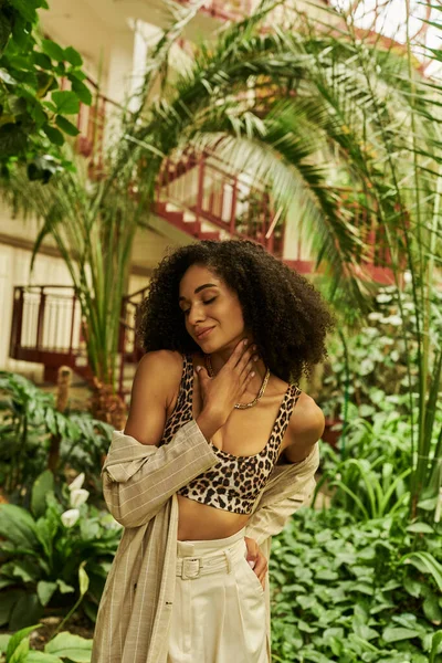 Pleased african american woman with curly hair in trendy look touching neck in tropical setting — Stock Photo