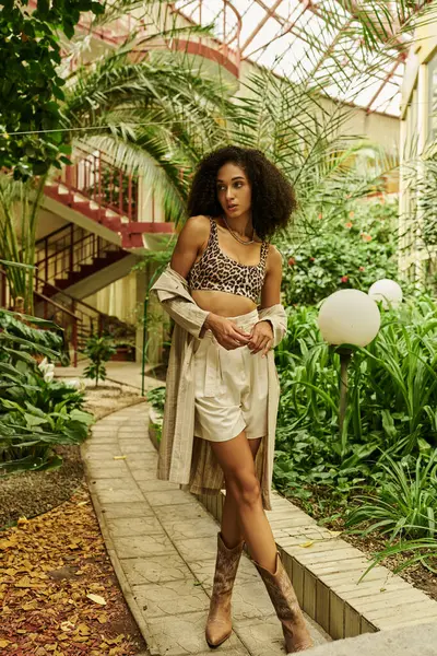 Femme afro-américaine élégante et jeune avec les cheveux bouclés debout dans un look branché dans un cadre tropical — Photo de stock