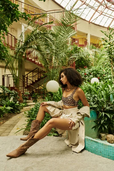 Longitud completa de la joven mujer negra con el pelo rizado posando en su aspecto de moda en un entorno botánico - foto de stock