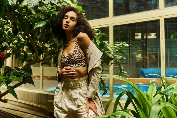 Кудрявая африканская американка позирует в своем модном облике в экзотической зеленой обстановке с тропическими растениями — стоковое фото