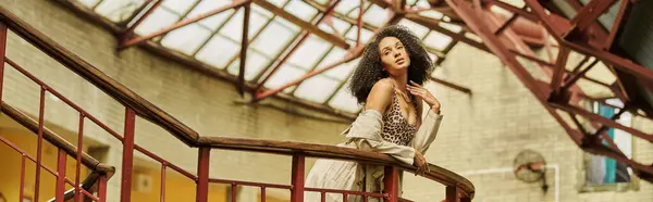 Curly chic african american woman in animal print look posing against industrial backdrop, banner — Stock Photo