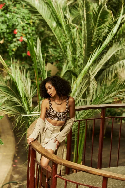 Jeune femme noire à la mode avec des cheveux bouclés posant sur un pont métallique dans un cadre de jardin luxuriant — Photo de stock