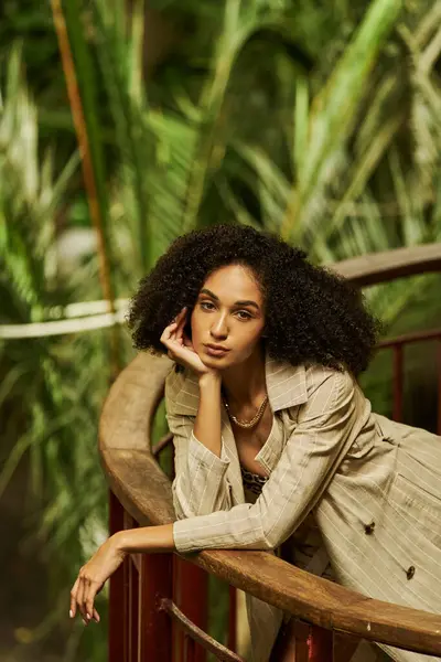 Fashionable young black woman with curly hair leaning on metallic structure in green garden setting — Stock Photo