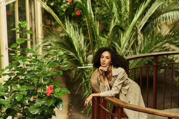 Stylish young black woman with curly hair leaning on metallic structure in green garden setting — Stock Photo