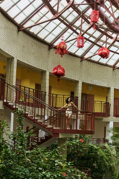 Look from afar, curly young black woman in animal print, surrounded by greenery and red lanterns — Stock Photo