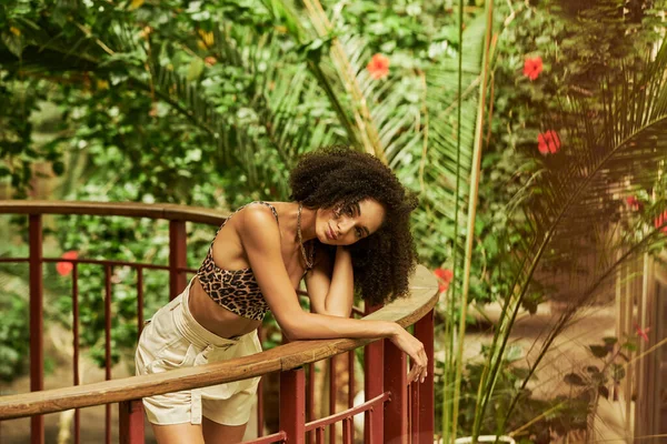 Flirty african american woman with curly hair posing on metallic bridge in green botanical setting — Stock Photo