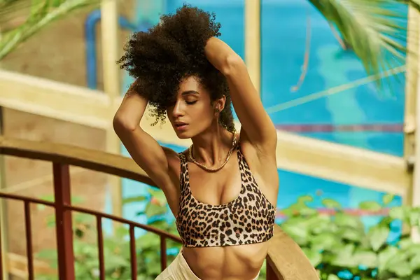 Elegante mujer afroamericana en estampado de leopardo mira levantando su cabello rizado y disfruta de tranquilidad - foto de stock