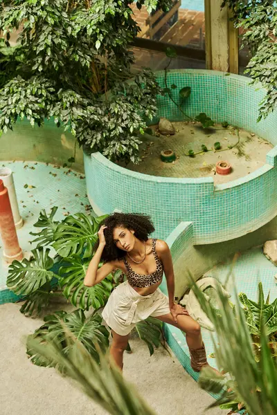 Trendy donna riccia africana americana in abbigliamento chic colpisce una posa in verdeggiante conservatorio — Foto stock