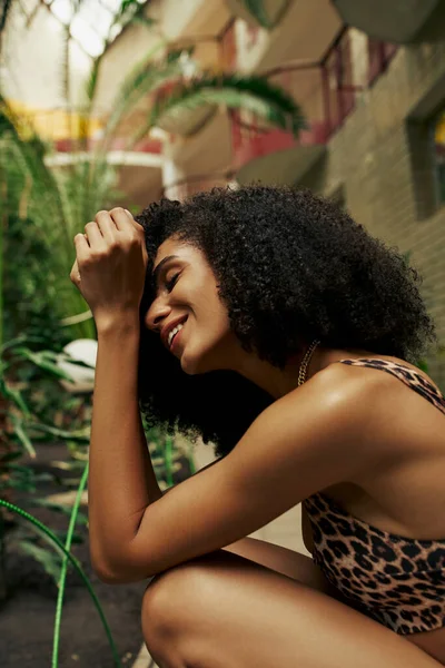 Fröhliche afrikanisch-amerikanische lockige Frau in schicker Kleidung, die lächelt und um grüne Pflanzen posiert — Stockfoto