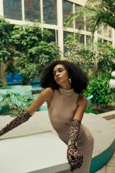 Alluring african american woman in dress and animal print gloves posing among plants in urban garden — Stock Photo