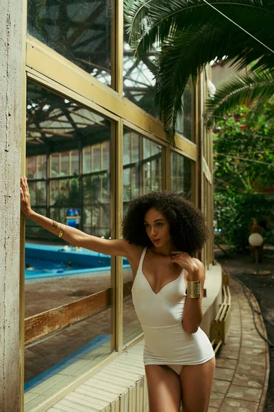 Кудрявая африканская американка в купальнике позирует в зеленом и экзотическом городском саду, разнообразие — стоковое фото