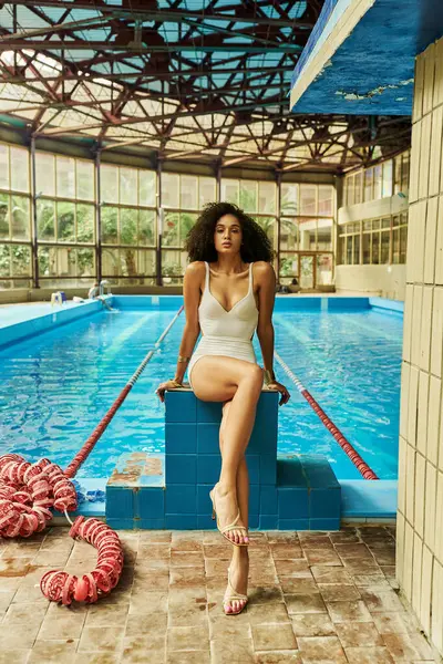 Lockige und hübsche Afroamerikanerin in eleganter Badebekleidung und High Heels am Pool sitzend — Stockfoto