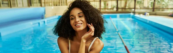 Feliz mulher afro-americana em roupa de banho sentado à beira da piscina com água azul, bandeira — Fotografia de Stock