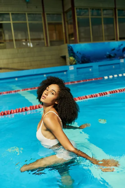 Young and curly african american woman in bathing suit standing by pool ladder with blue water — Stock Photo