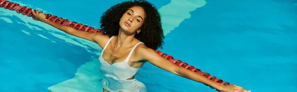 Jovem e encaracolado mulher americana africana nadando na água azul da piscina e olhando para a câmera, bandeira — Fotografia de Stock